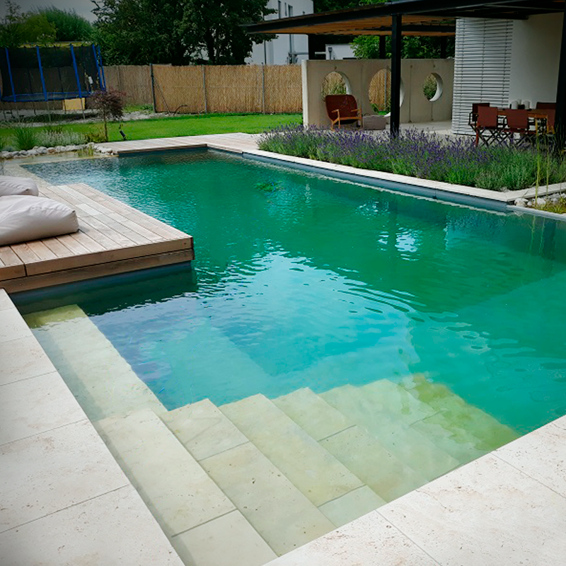 Dynamische Gartengestaltung Eder Naturpool Blick auf ein wunderschönes Naturpool mit glasklarem Wasser, Einstiegstreppe, seitlichem Holzsteg auf dem man 2 Gartenliegen sieht. Im Hintergrund ein Blumenbett mit einer gemütlichen Sitzecke.
