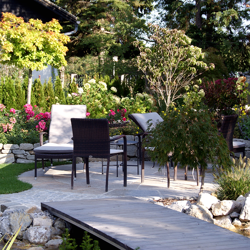 Dynamische Gartengestaltung Eder Garten Blick auf eine gemütliche Sitzecke im Garten. Im Vordergrund sieht man einen Holzsteg. Im Hintergrund eine kleine Steinmauer mit Ziersträuchern.