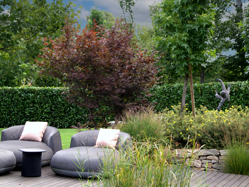 Dynamische Gartengestaltung Eder Gartenanlage Gemütliche Sofaecke auf einer Holzterrasse in einem einladenden Garten. Im Hintergrund sieht man Sträucher und Bäume.