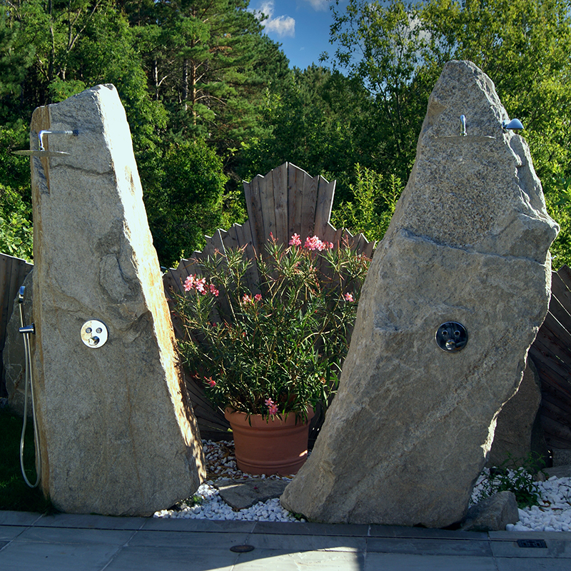 Dynamische Gartengestaltung Eder Gartendusche Gartendusche aus Natursteinen. Im Hintergrund Bäume in sattem grün und strahlend blauer Himmel.