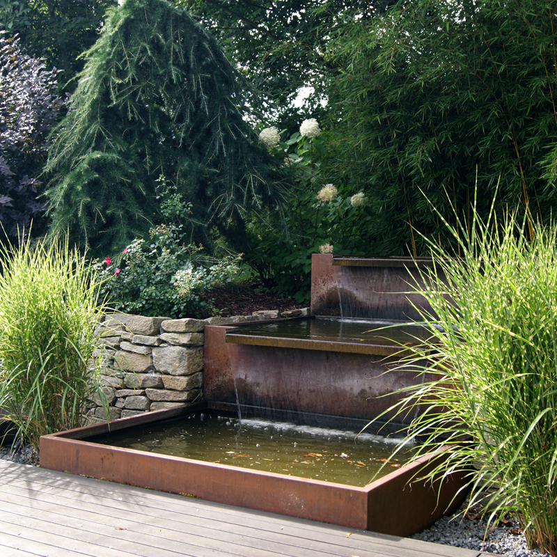 Dynamische Gartengestaltung Eder Gartenanlage Wasserlauf umrandet von Hochbeeten aus Natursteinen, seitlich sind Ziergräser gepflanzt, im Vordergrund sieht man eine Holzterrasse.