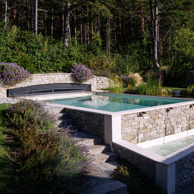 Dynamische Gartengestaltung Eder Naturpool Naturpool in Mitten einer idyllischen Gartenlandschaft, eingebettet in eine dekorative Mauer aus Natursteinen.