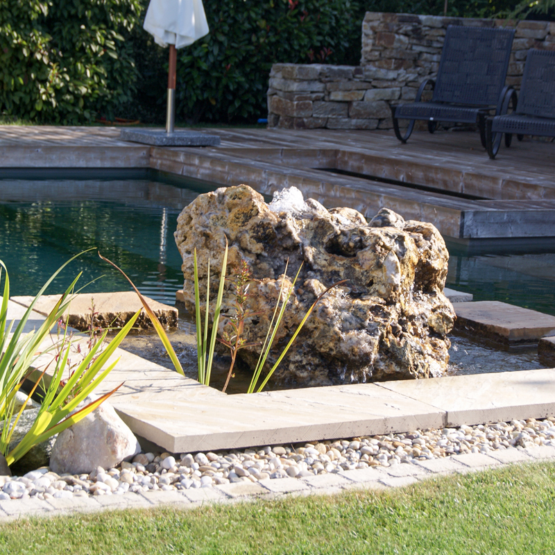 Dynamische Gartengestaltung Eder Naturpool Naturpool mit glasklarem Wasser, den vorderen Bereich ziert ein mit Steinplatten umrahmter Quellstein. Im Hintergrund eine gemütliche Holzterrasse mit zwei Liegestühlen.